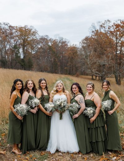 Bridal party consisting of eight women in olive green dresses, holding bouquets of white flowers, with one woman in a white wedding gown.