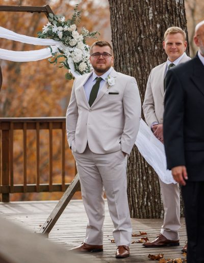 The groom in a light-colored suit with a green tie standing at a wedding venue, with a floral arch in the background.