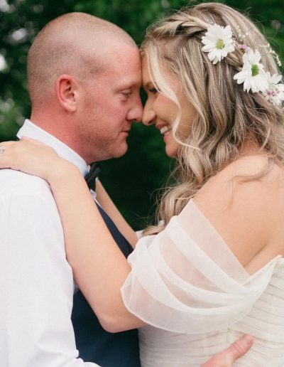 Couple embracing with foreheads touching, the woman adorned with flowers in her hair and wearing a white dress, while the man is in a formal outfit.