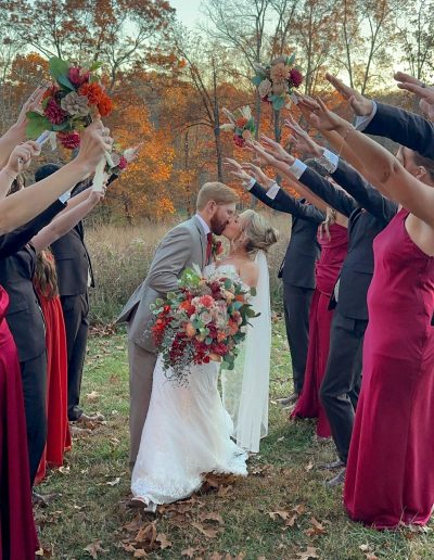 Bride and groom kissing under an arch of hands held by wedding party members, surrounded by autumn foliage.