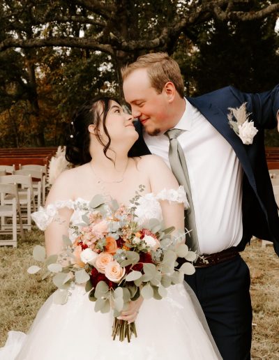 Bride and groom sharing a moment outdoors, with the bride holding a bouquet of flowers and the groom in a suit, both smiling at each other.