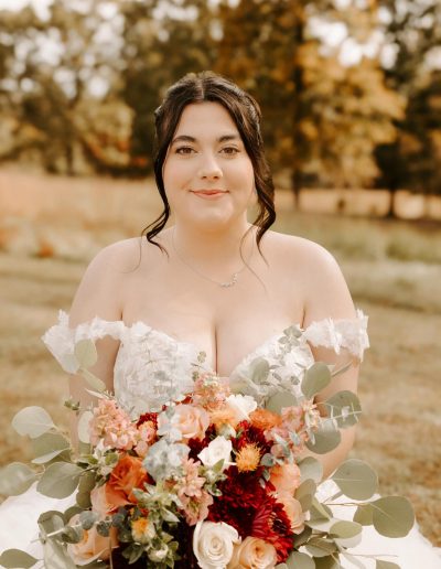 Bride holding a bouquet of flowers, wearing a strapless wedding gown with lace details, standing outdoors with a natural background.