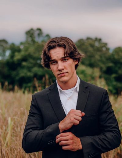 Young man in a black suit adjusting his cuff while standing in a field with tall grass.