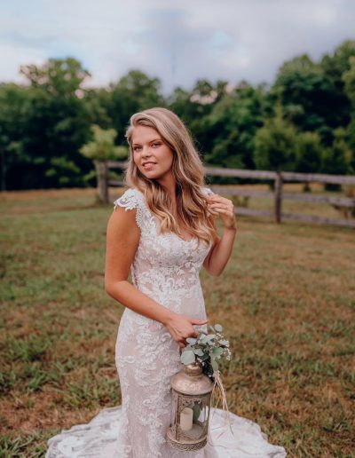 Bride in a lace wedding dress holding a lantern and greenery, standing in a grassy area with trees in the background.