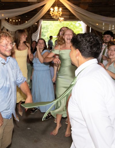 Wedding guests dance beneath draped fabric and string lights at a rustic venue during a lively reception celebration.