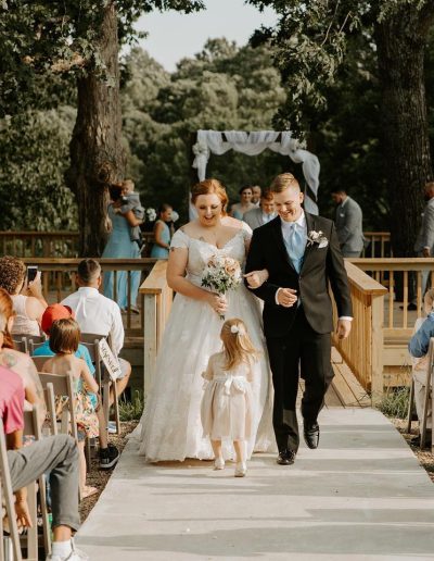 Bride and groom walk down a wooden aisle beneath a white draped arch at an outdoor wooded wedding ceremony with seated guests in warm sunlight.