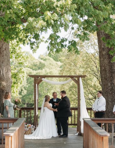 Outdoor wedding ceremony beneath tall trees with a wooden arch, white drapery, and a couple exchanging vows on a rustic deck at Heartland Meadows.