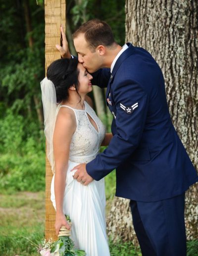 A bride in a white wedding dress kisses her groom in a blue military uniform beneath tall trees outdoors.
