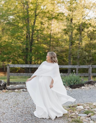 A bride in a flowing white gown smiles on a gravel path at Heartland Meadows, framed by a rustic fence, stone border, and green trees.