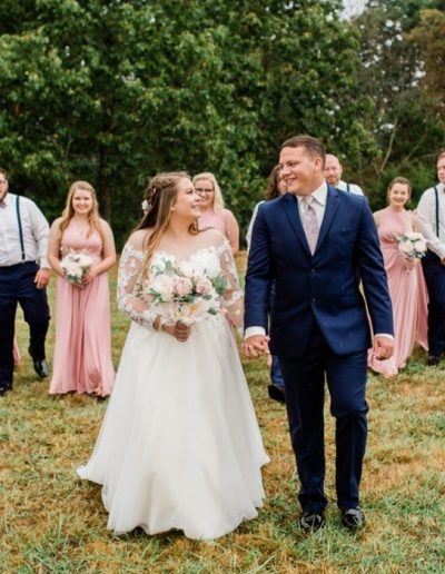 Newlyweds walk hand in hand across Heartland Meadows' lush green lawn, surrounded by bridesmaids in pink dresses and groomsmen in navy and white.