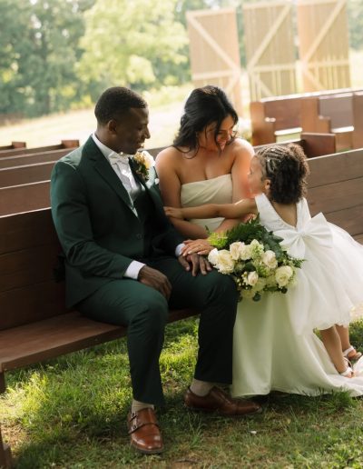 A groom in a dark green suit sits beside a bride in a strapless gown while a little girl in white leans in with flowers.