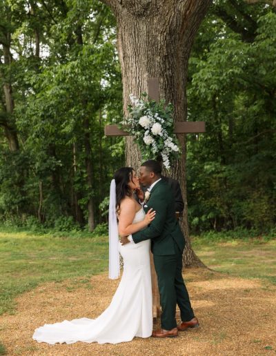 A bride in a white gown and veil kisses a groom in a dark green suit beneath a floral wooden cross.