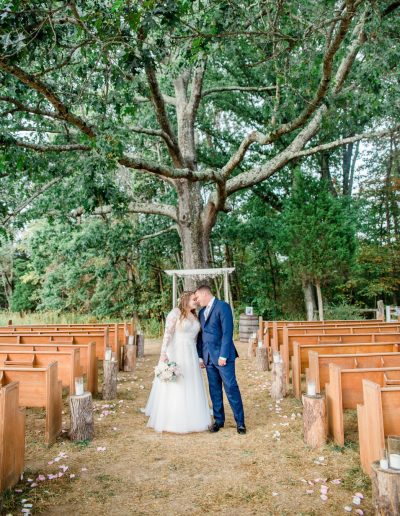 Bride and groom share a quiet kiss beneath a sprawling oak at Heartland Meadows, with wooden pews, scattered petals, and lush greenery all around.