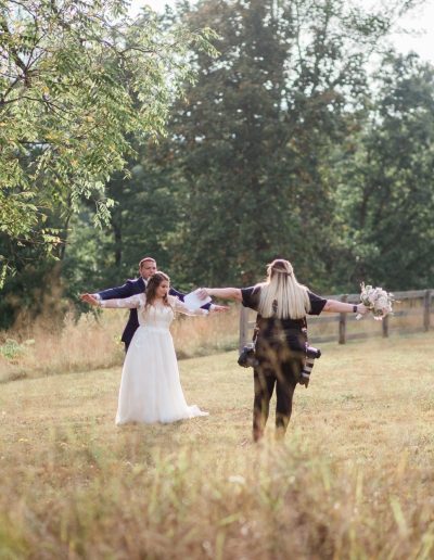At Heartland Meadows, a bride in a white lace gown and groom stand with arms outstretched while a photographer directs them in a sunlit meadow.