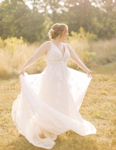 A bride in a white lace wedding dress lifts her flowing skirt in a sunlit meadow surrounded by tall grasses and trees.