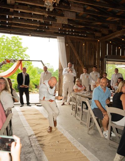 Wedding guests gather in a rustic barn as a groom dances down the aisle beneath a chandelier and floral drapery.