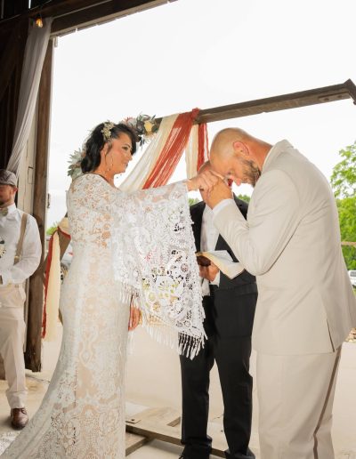 At Heartland Meadows, a bride in a lace gown bows beside her groom under a rustic wedding arbor during the ceremony.