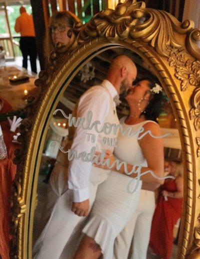 A bride and groom kiss in an ornate gold mirror, with “Welcome to our wedding” handwritten across the glass.
