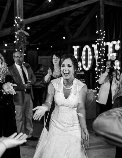 Black and white wedding reception with a laughing bride in a lace gown dancing among guests, string lights, and a glowing LOVE sign indoors warmly.