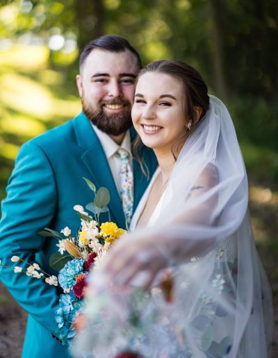 Smiling newlywed couple outdoors, with the groom in a teal suit and the bride holding a colorful bouquet beneath her flowing veil