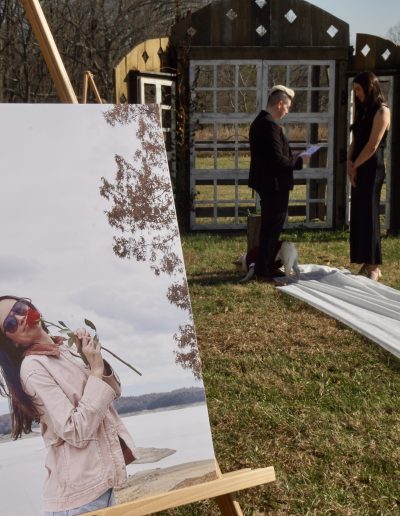 A large portrait of a woman holding a rose on display, with a couple standing in front of a wedding altar outdoors.