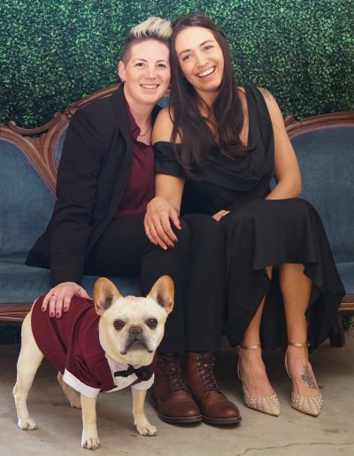 A couple seated on a blue couch, one wearing a black dress and the other in a suit, with a French Bulldog in a maroon tuxedo in front.