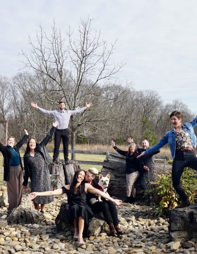 Group of people posing outdoors on rocks and logs, with some standing and others seated, against a backdrop of trees and a cloudy sky.