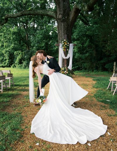 A bride and groom kiss beneath a large oak tree, framed by white drapery and flowers along a wooded outdoor wedding aisle.