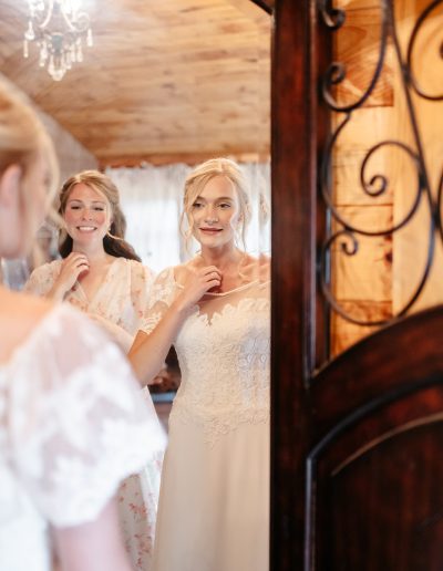 A bride adjusts a pearl necklace in a mirror while a bridesmaid smiles beside her in a warm wood-paneled room.
