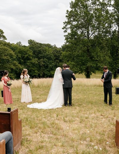 A bride and groom stand at an outdoor wedding ceremony in a grassy meadow surrounded by bridesmaids, groomsmen, and guests.
