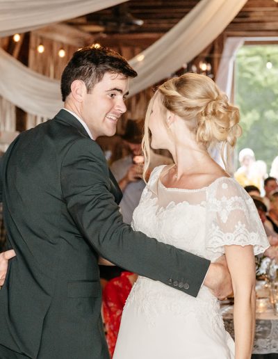 A bride in a white lace gown and a groom in a dark suit share a dance at a rustic wedding reception.