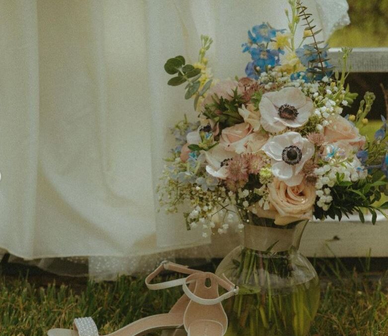 A pair of bridal shoes next to a vase filled with a floral bouquet on grass.